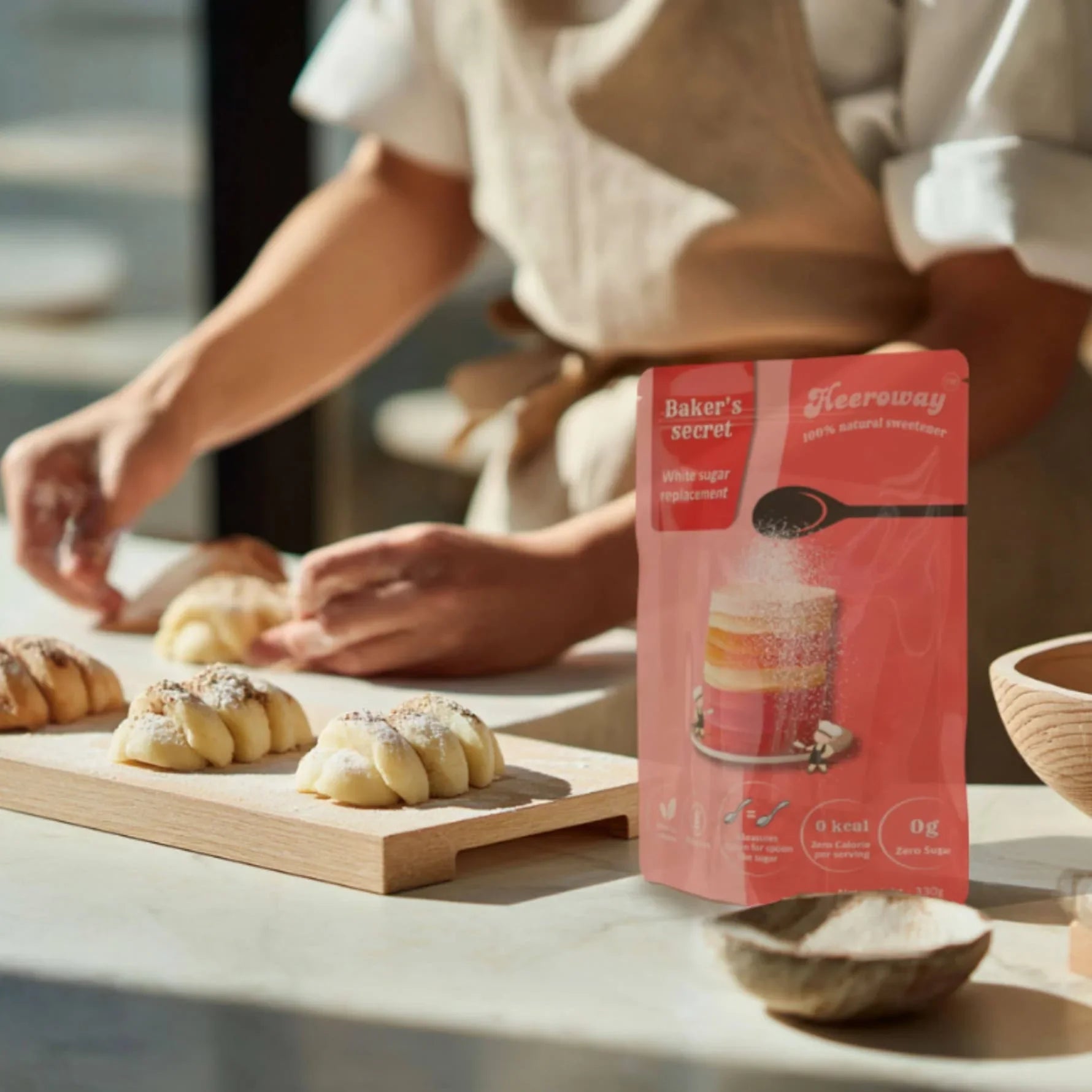 Heeroway Baker's Secret white sugar replacement pouch on a kitchen counter while a baker prepares and sprinkles sugar over freshly made bread rolls for product page.