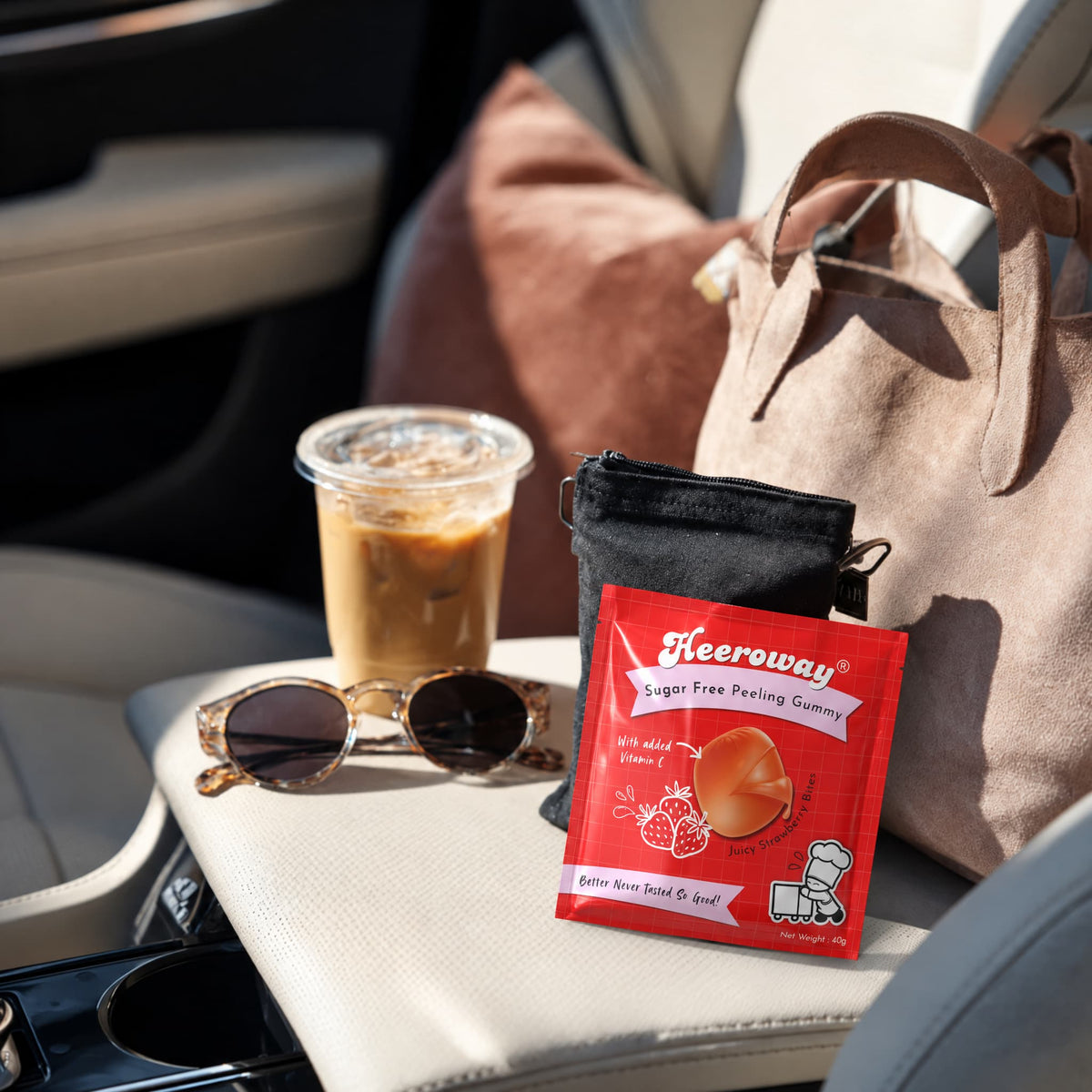 A packet of Heeroway Strawberry Sugar Free Peeling Gummies placed on a beige car seat next to a clear cup of iced coffee, tortoiseshell sunglasses, a black pouch, and a beige tote bag.