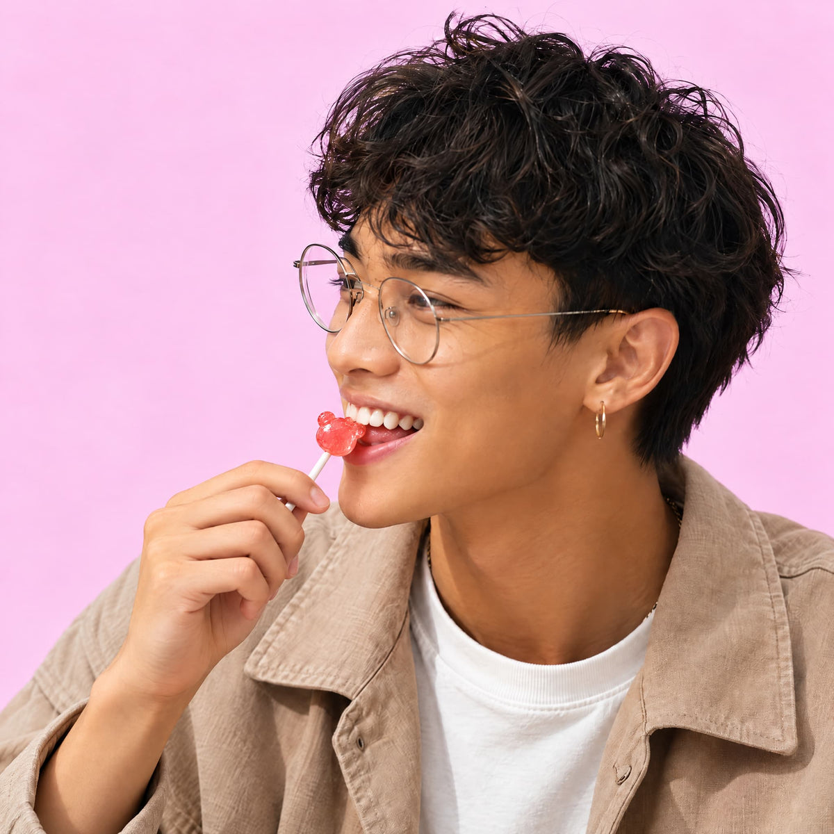 A man with curly dark hair and round metal-framed glasses against a solid pink background, holding a red bear-shaped lollipop. 

