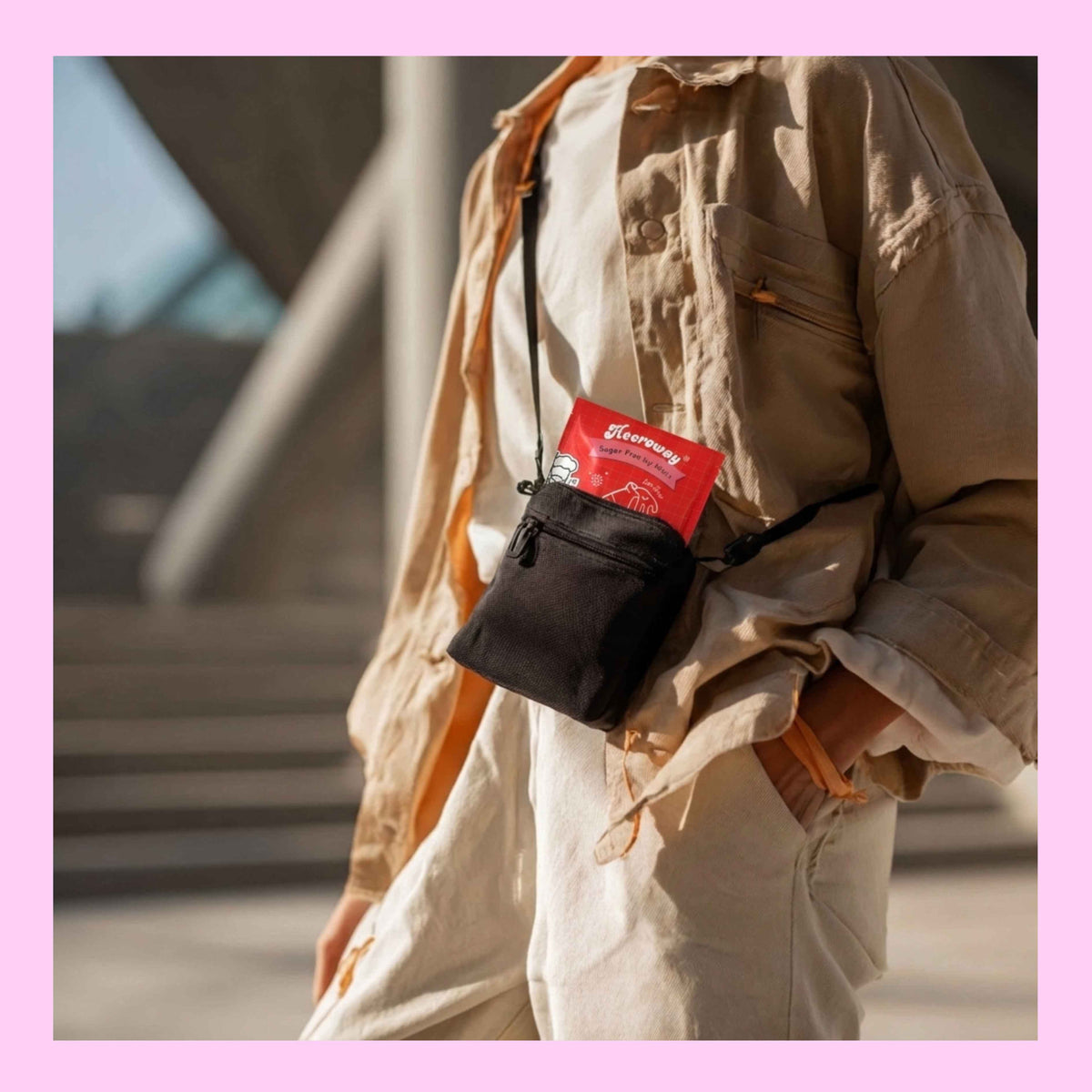 A person wearing a beige shirt and pants carries a small black crossbody bag with Heeroway cola mint tucked inside, photographed outdoors with a blurred architectural background.