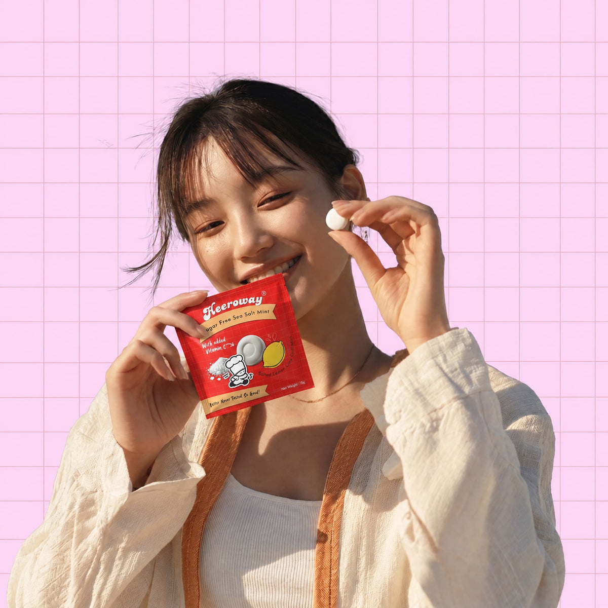 A smiling woman holds a small red packet of Heeroway lemon sea salt mint  set against a pink grid background.