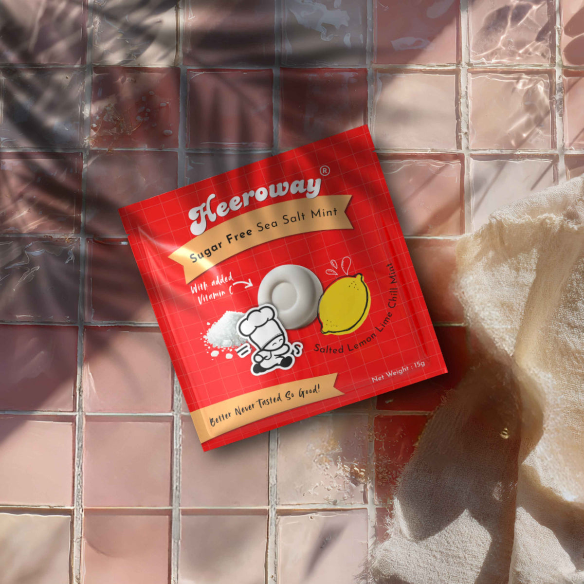 A red packet of Heeroway Sugar Free Sea Salt Mint lemon flavor lies on a glossy pink tiled surface with a beige towel nearby.