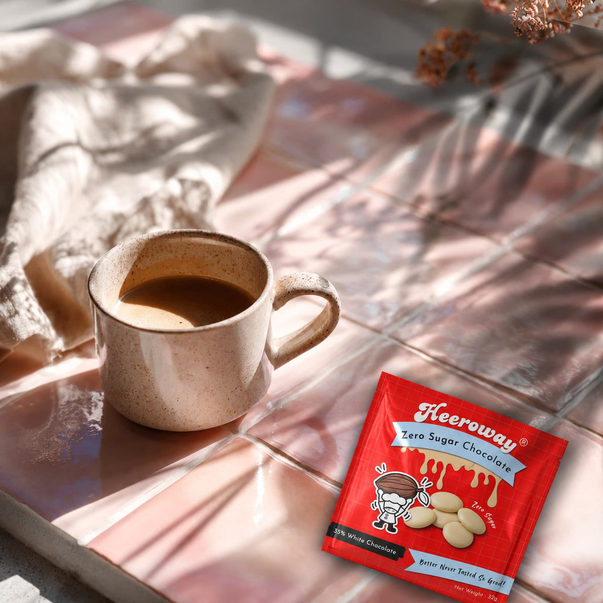 A packet of Heeroway White Chocolate lies on a pink tiled surface next to a ceramic mug filled with coffee, with warm sunlight casting shadows across the scene.