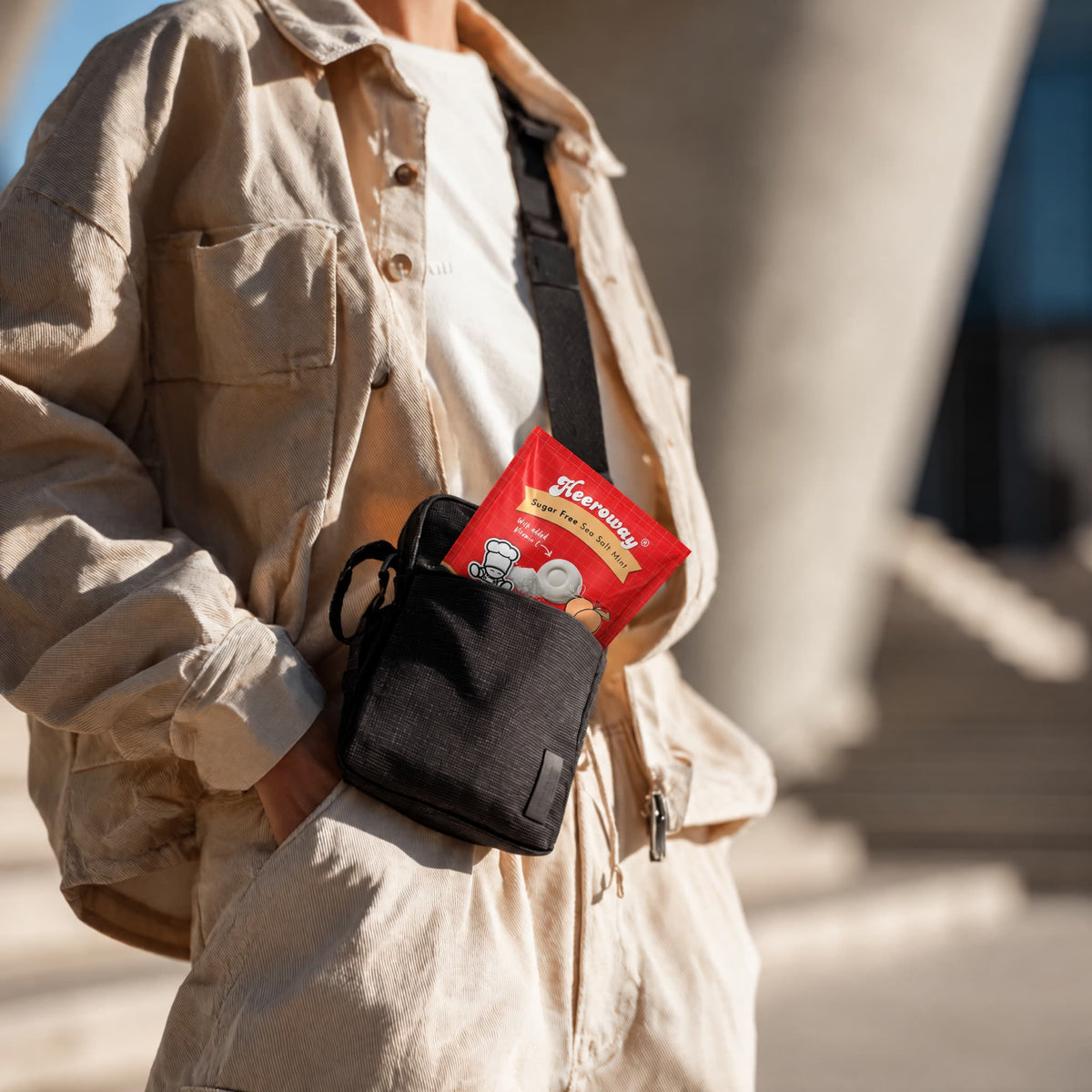 A person outdoors wearing a beige overshirt and matching pants, carrying a small black crossbody pouch at the waist. The pouch contains a packet of Heeroway peach sea salt mint  and the background shows blurred stone architecture in bright sunlight.
