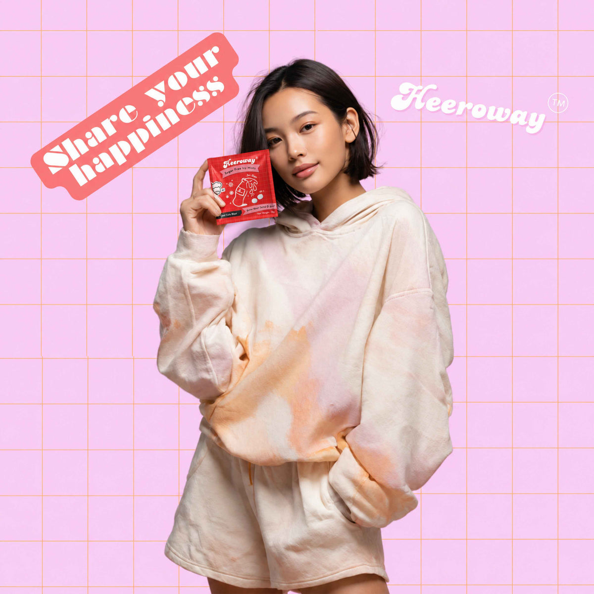 A woman wearing a pastel tie-dye hoodie and shorts holds up a packet of Heeroway cola mint  against a pink grid background with 'Share your happiness' and 'Heeroway' text overlays.