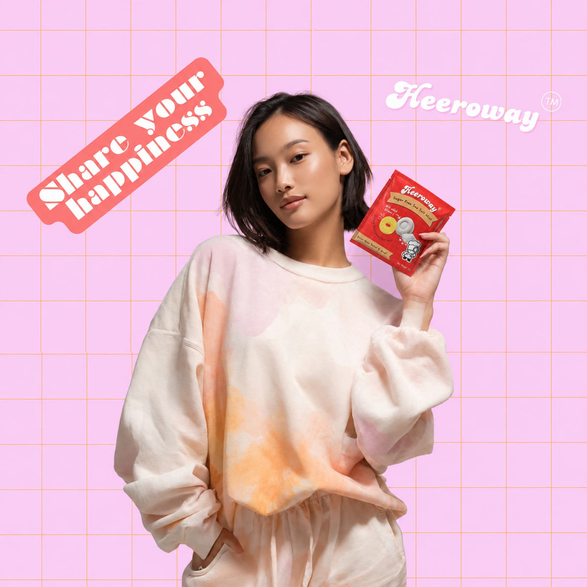 A woman wearing a pastel tie-dye sweatshirt and matching pants holds a packet of Heeroway pineapple sea salt mint pouch  against a pink grid background with 'Share your happiness' and 'Heeroway' text overlays.