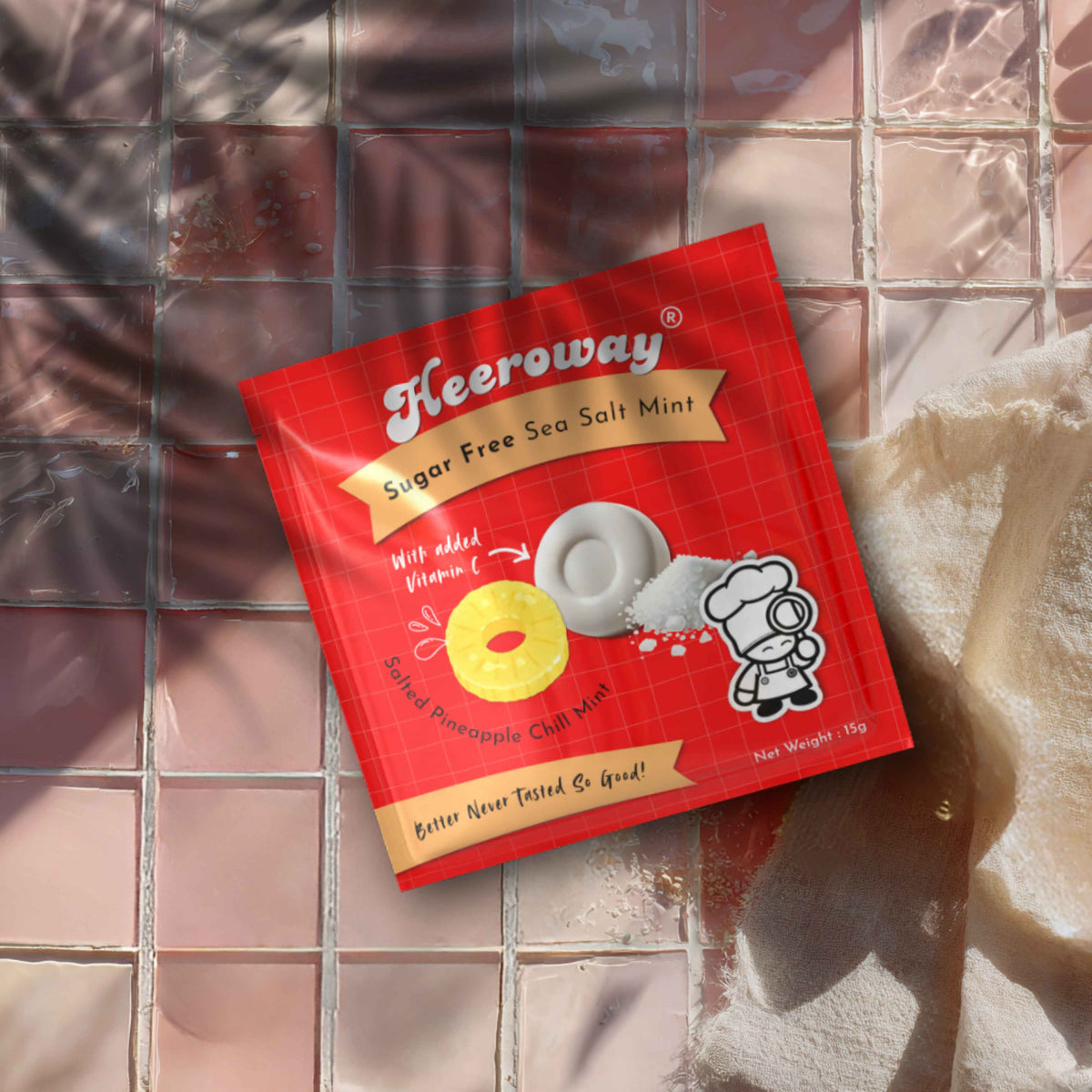 A packet of Heeroway pineapple Sugar Free Sea Salt Mint candy lies on a glossy pink tiled surface with a beige cloth partially visible on the right.
